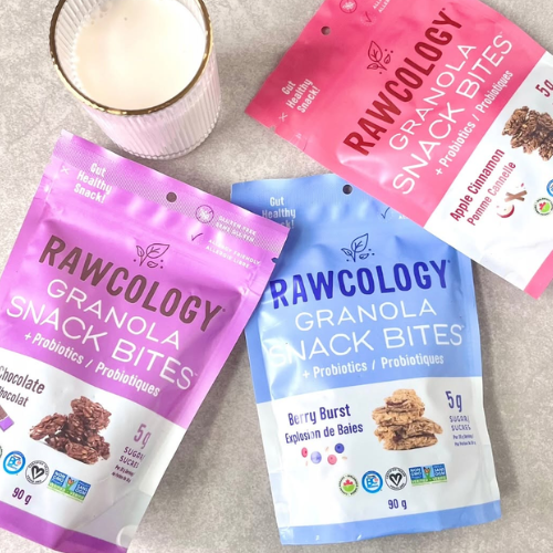 Three RAWCOLOGY granola snack bites pouches arranged on a light stone surface with a white cup of milk. The pouches are pink, blue, and purple, featuring the RAWCOLOGY brand name.
