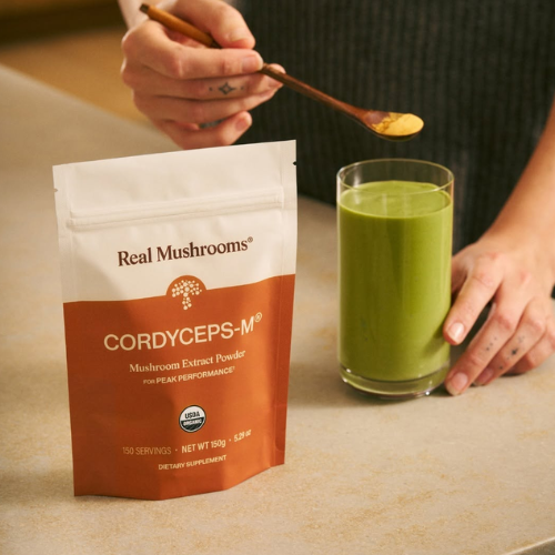 Real Mushrooms Cordyceps-M powder package with a glass of green liquid on a table.