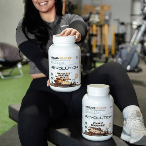 Person holding two 'Vegan Smart Revolution' protein powder containers in a gym setting