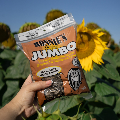 Hand holding a package of Ronnie's Original Jumbo Roasted Sunflower Seeds in front of sunflowers.
