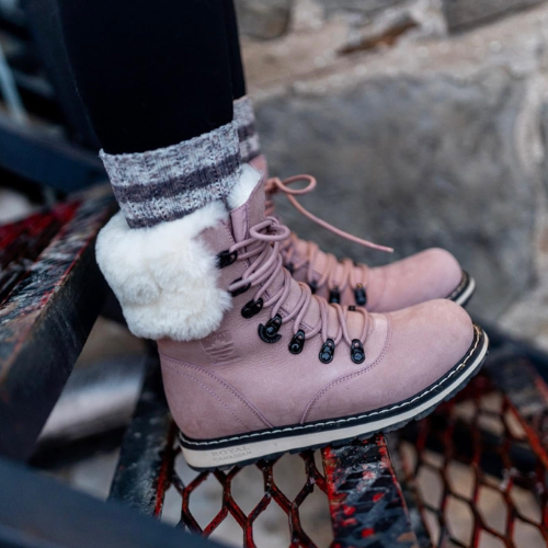 A close-up of a person wearing dusty pink suede lace-up boots with a fluffy white faux-fur trim and black eyelets, standing on a metal grate.
