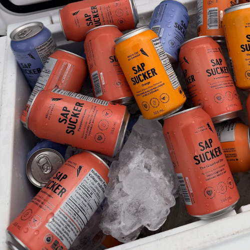 Multiple SAP SUCKER energy drink cans in orange and blue packaging, some with condensation, arranged in a white cooler tray with ice.
