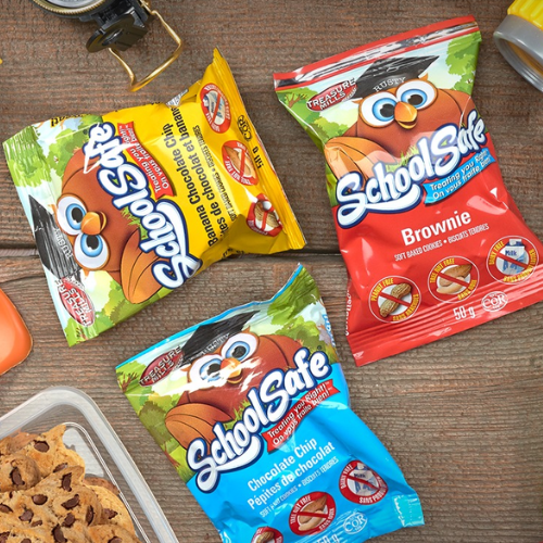 Three colourful packages of 'School Safe' snacks on a wooden surface with cookies.
