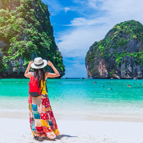 Woman in a colourful dress and sun hat standing on a tropical beach with limestone cliffs and clear blue water.