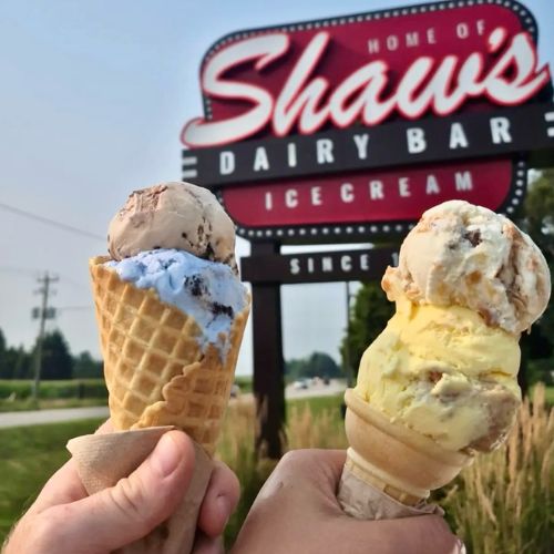 Two ice cream cones held up in front of Shaw's Dairy Bar sign.