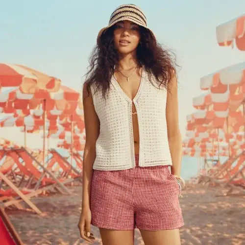 Woman in Simons summer outfit with red gingham shorts and crochet vest standing on a beach with striped umbrellas