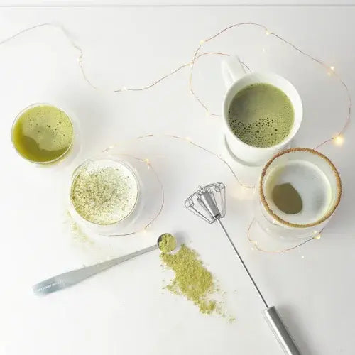 Matcha green tea in white cups with a whisk and powder on a light surface.
