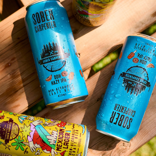 Two blue cans of 'Sober Carpenter' non-alcoholic craft beer on a wooden surface with a colorful can partially visible.