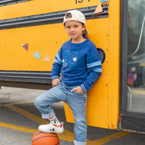 Child in blue Souris Mini sweatshirt with number 89 leaning against a yellow school bus