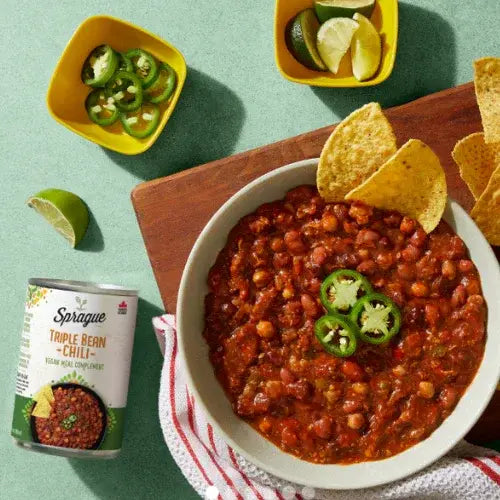 Bowl of chili with tortilla chips, jalapeños, and lime wedges on a green background with Sprague chili can.