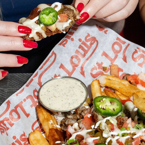 Person holding a loaded nacho with a side of fries and sauce on a St. Louis branded paper.