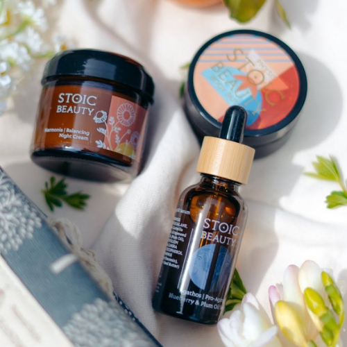 Stoic Beauty skincare products including a jar, a dropper bottle, and a round container on a floral background.