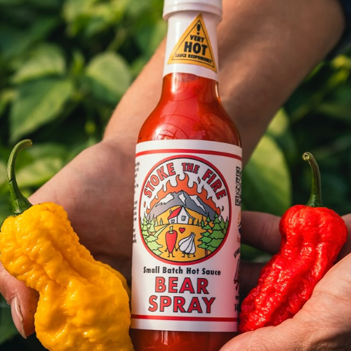 Bottle of Bear Spray hot sauce held between two peppers with a blurred green background