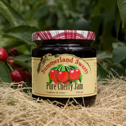 Jar of Summerland Sweet Pure Cherry Jam with a red and white checkered lid, surrounded by cherries and greenery.