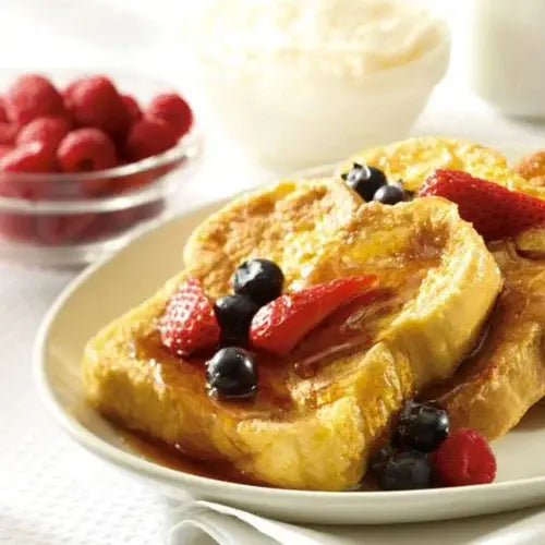 French toast topped with fresh strawberries, blueberries, and raspberries, drizzled with syrup, served on a cream-coloured plate with a side bowl of raspberries and a dish of whipped cream in the background.