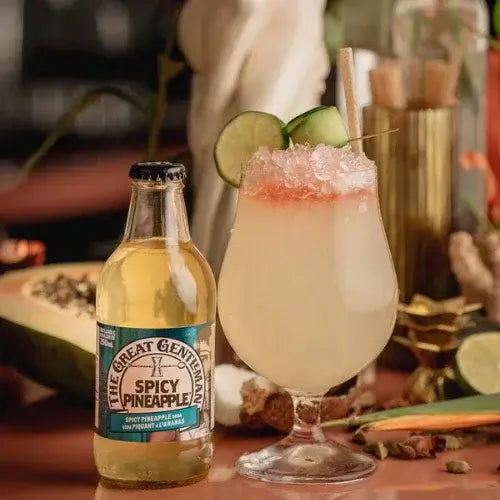 Bottle of 'The Great Gentleman Spicy Pineapple' next to a cocktail with lime garnish on a wooden surface.