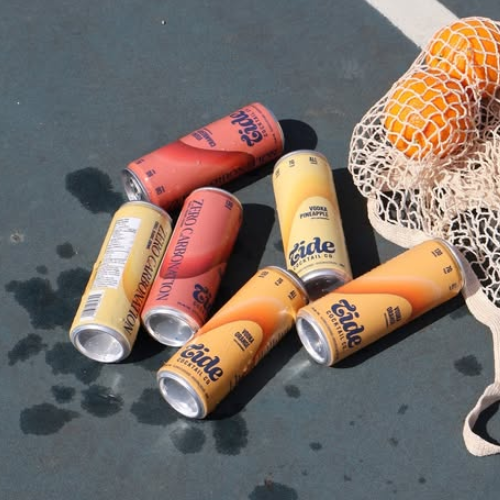 Cans of Tide Cocktail Co. drinks on a dark surface with a bag of oranges in the background