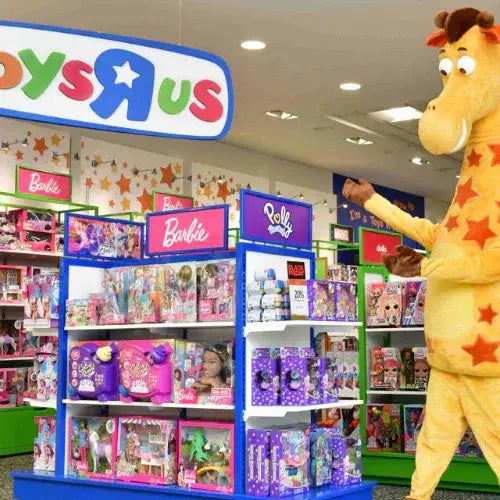 Inside a Toys 'R' Us store, a large giraffe mascot (Geoffrey the Giraffe) gestures toward colourful toy displays featuring Barbie and Polly Pocket products. The store is bright and cheerful with well-stocked shelves and playful signage.