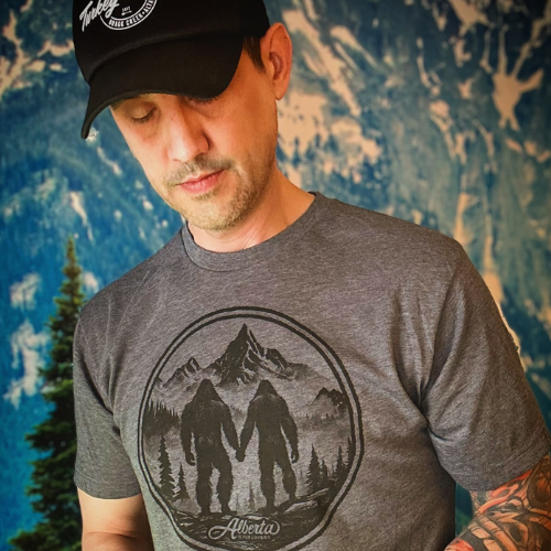 Man wearing a gray t-shirt with a Sasquatch graphic and a black cap against a textured blue background