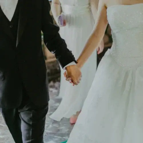 A bride and groom holding hands, walking together. The bride is wearing a strapless white wedding gown with delicate embellishments, while the groom is dressed in a classic black suit. A bridesmaid in a white dress is visible in the background