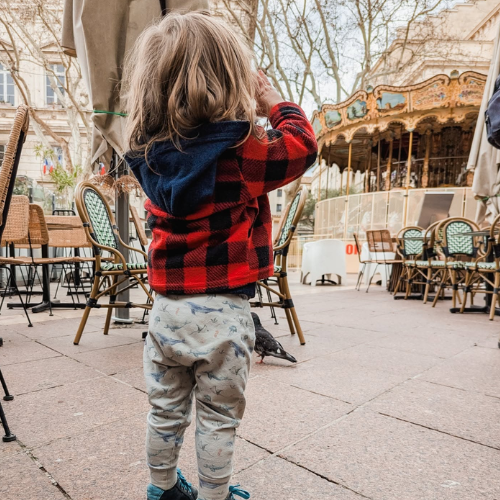 A toddler standing in a cafe patio area, viewed from behind, wearing a red and black buffalo plaid hooded sweatshirt with a navy blue hood, and light gray patterned pants.