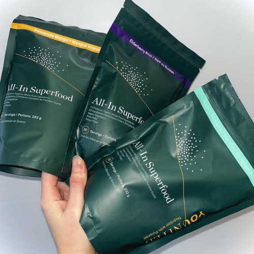 Three 'All-In Superfood' packages held by a hand on a light gray background